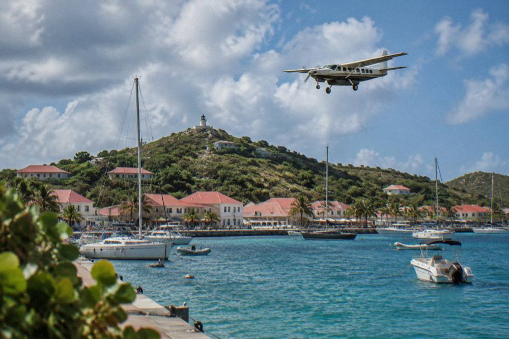 A single-engine turboprop flying low over green Caribbean hills toward a small island airport