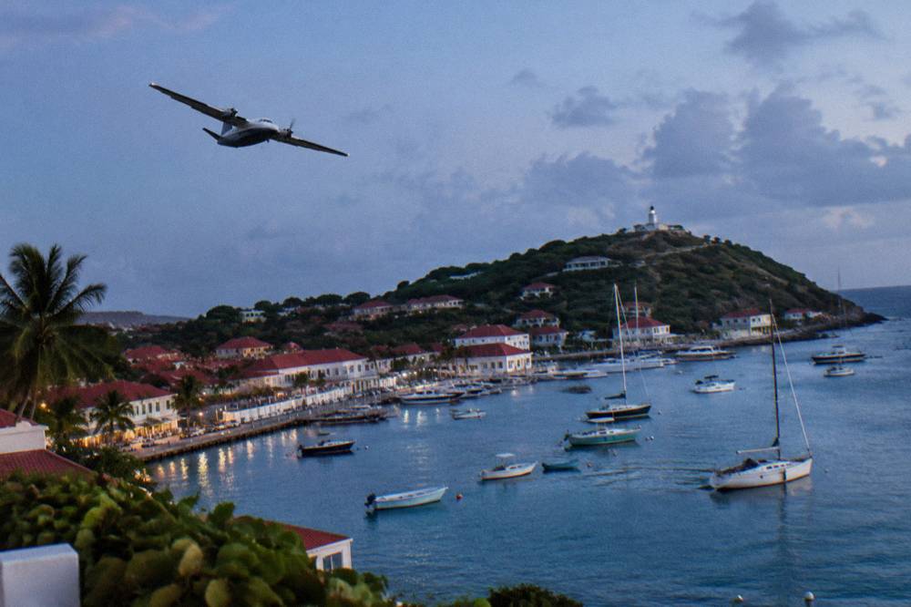 A twin-engine turboprop banking over a Caribbean hillside with a harbor town visible below