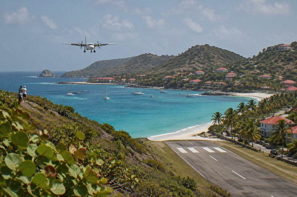 A short runway ending beside a palm-lined beach road with a bay and hills beyond