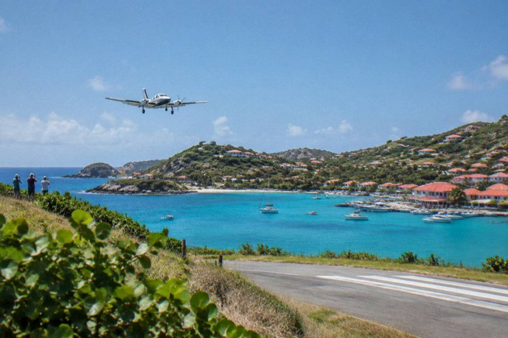 A small aircraft taking off from a short runway over turquoise Caribbean water at morning light
