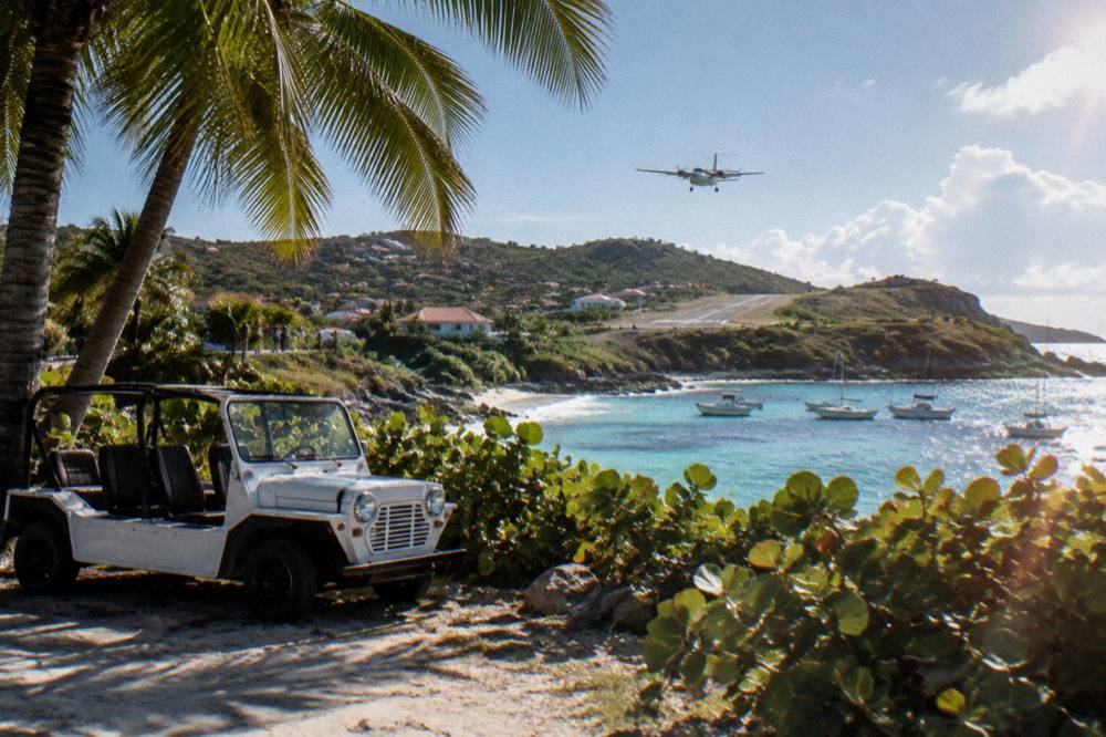 An open-air Mini Moke car parked beside palm trees near a Caribbean beach