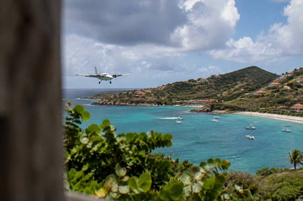 A small airplane flying low over turquoise Caribbean water toward a green island