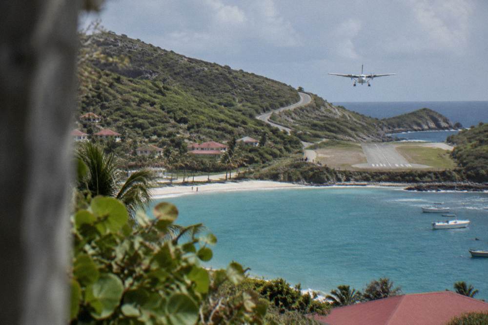A steep green hillside with a road winding down toward a short runway and a curved bay