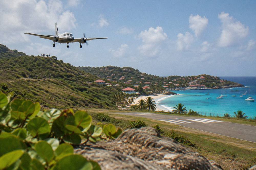 An aircraft descending low over a green hillside toward a short runway beside a Caribbean beach