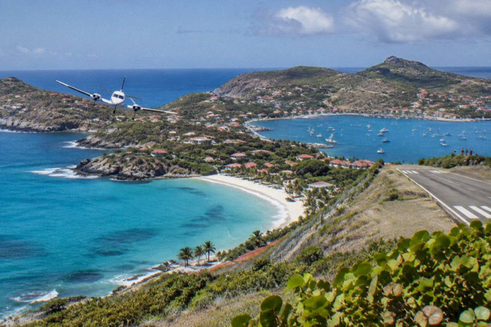 A light aircraft banking over a turquoise bay with white sand and green headlands