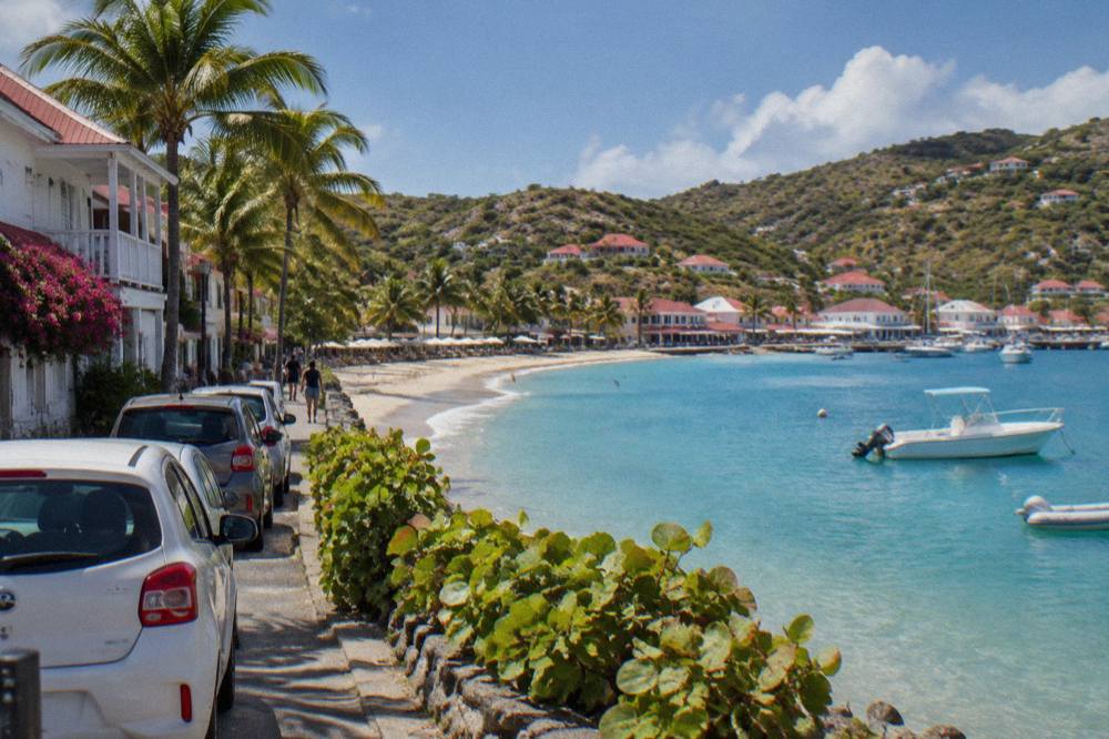 Small parked rental cars lined along a street of low white buildings in a Caribbean harbour town
