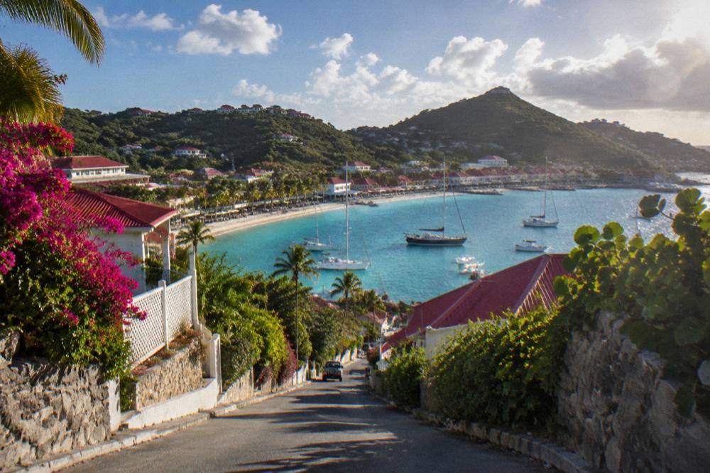 A driver's view down a steep hill toward a small Caribbean harbour with moored sailboats