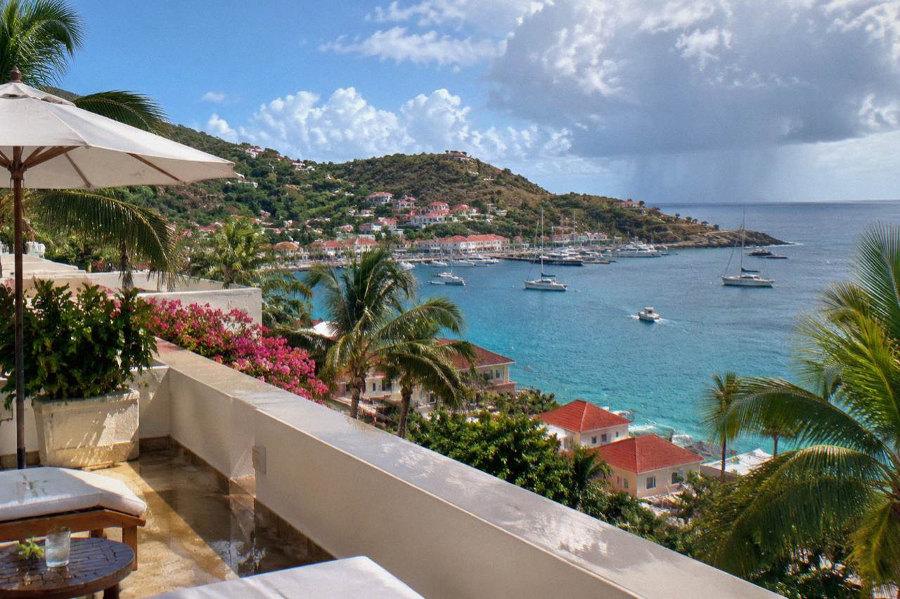 A cream-colored hotel terrace above the coastline of St Barts with palm trees and blue Caribbean water below