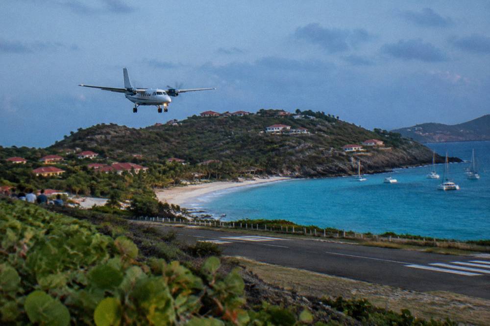 A small twin-engine turboprop airplane on a short runway beside a Caribbean beach with hills behind