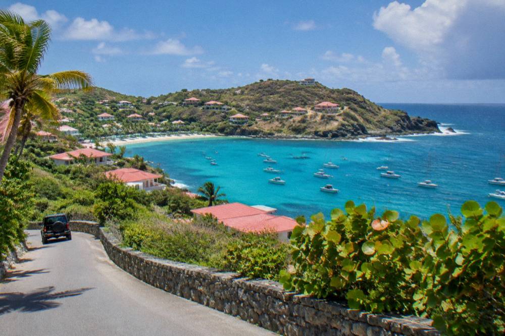 A hillside road curving past low villas with red roofs above a turquoise bay in the Caribbean