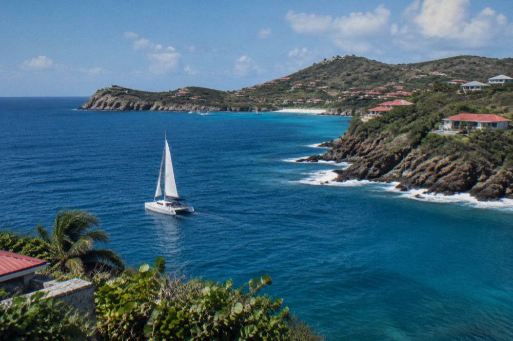 A sailing catamaran on open blue water approaching a green Caribbean island headland