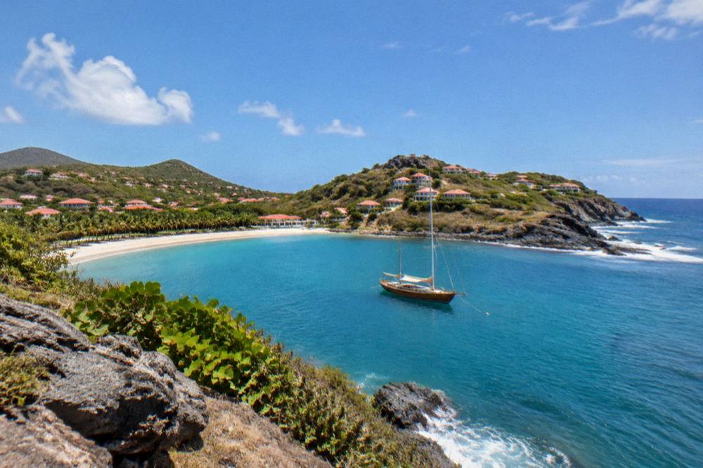 A wooden sailing boat anchored in a quiet bay with palm trees and a sandy beach behind it