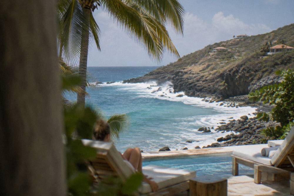 A hotel swimming pool terrace under palm trees with wooden loungers facing the ocean