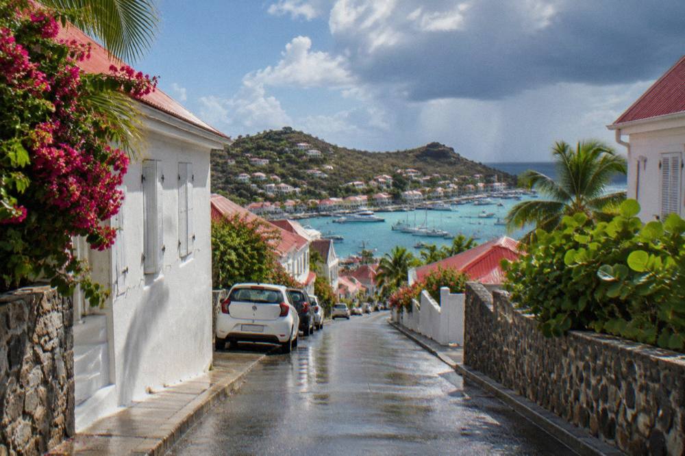 A narrow street in a Caribbean harbor town with low white buildings and parked small cars