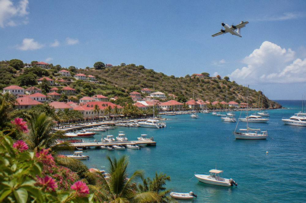 A twin-engine turboprop aircraft banking over green hills above blue Caribbean water near St Barts