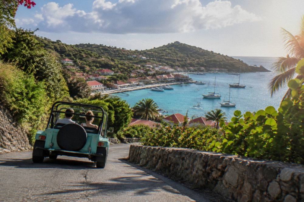 A small open-top Mini Moke car driving a narrow winding road between green hills in St Barts