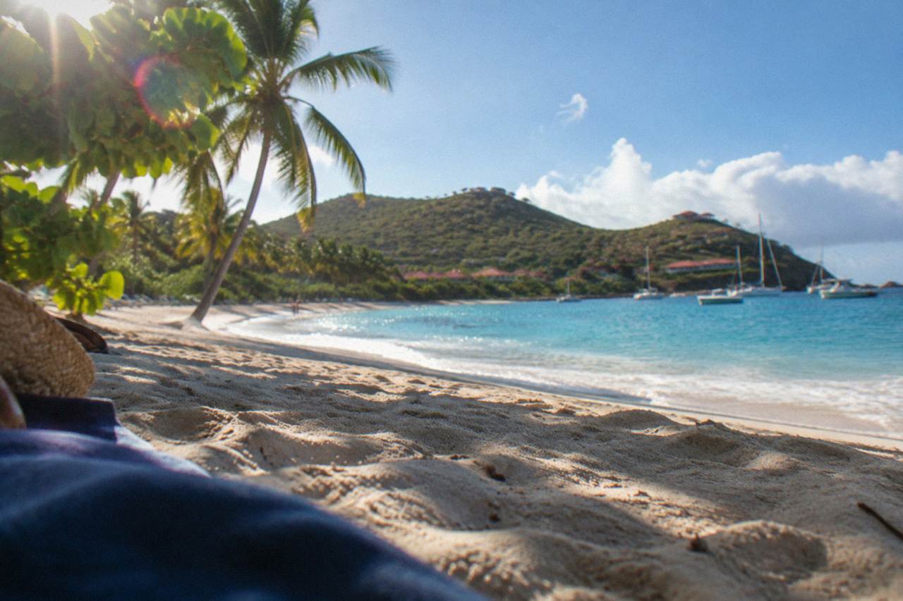 A white-sand beach with calm turquoise water and green hills behind it on the coast of St Barts
