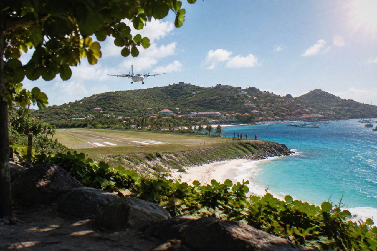 A small turboprop airplane on a short runway beside a Caribbean beach with hills behind it