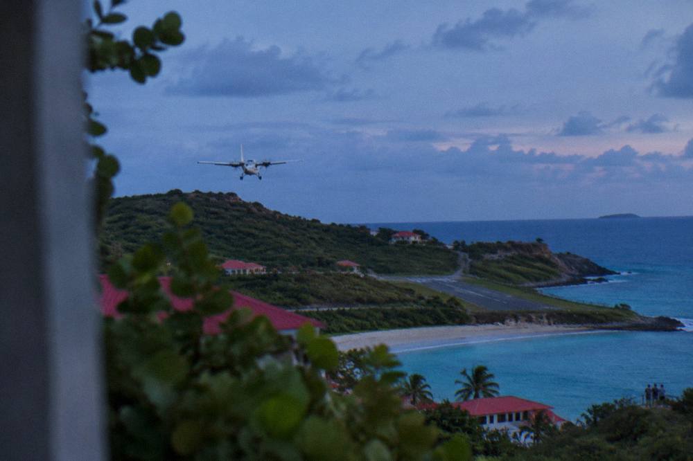 An airplane descending low over a green hill toward a short runway by turquoise sea