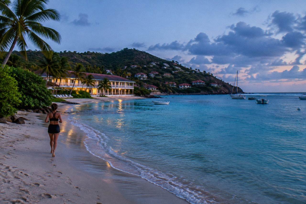 A low beachfront hotel building beside a calm turquoise bay with a sandy shore and green hills behind