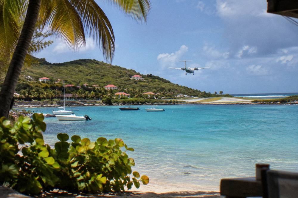 A small twin-engine turboprop aircraft flying low over a green hillside toward a short seaside runway