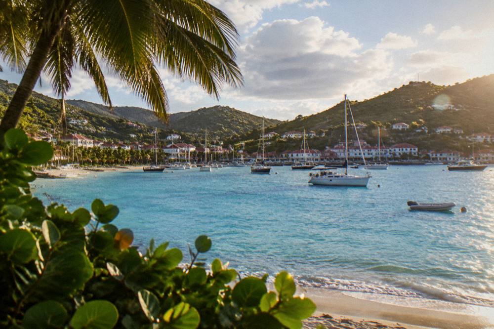 An evening view of a small harbour with white buildings, moored sailboats and hills in warm light