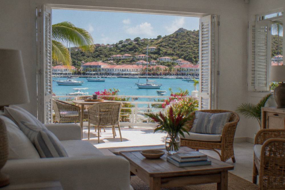 Open-plan rental apartment interior with white walls, rattan furniture and shutters open to a sea view