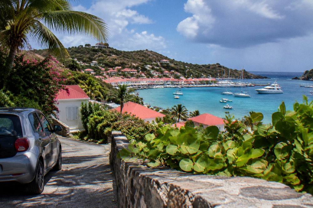 Small rental car parked on a narrow lane beside tropical vegetation and a glimpse of the sea