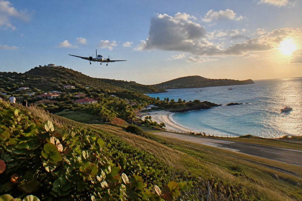 A light aircraft descending low over a green hillside toward a beachside runway