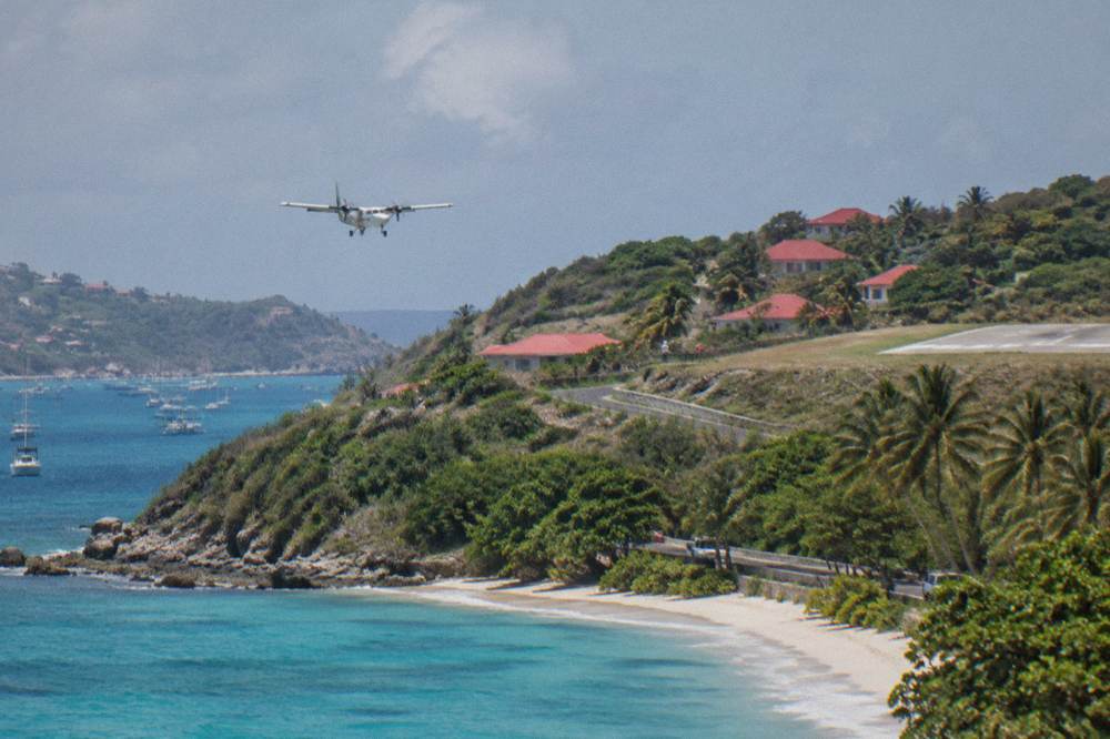 A small aircraft on final approach low over a green hill above a beach road