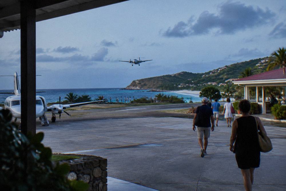 Passengers walking from a small aircraft across a warm airport apron toward a low terminal building