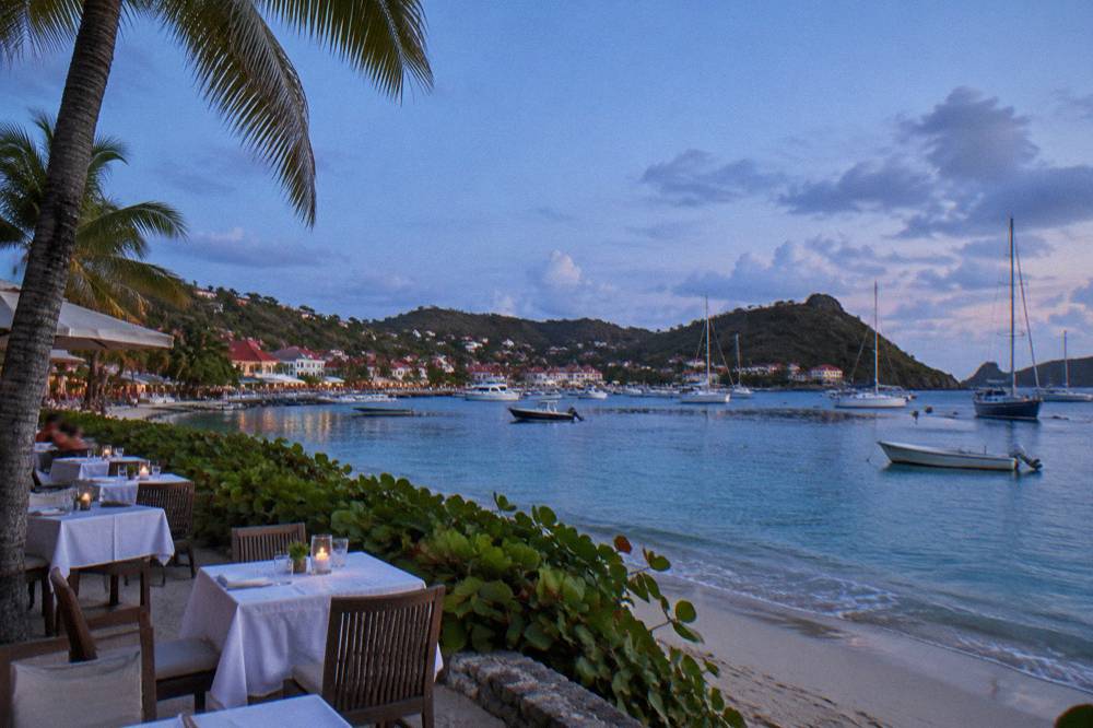 A beach restaurant terrace with white tablecloths beside palm trees and calm sea