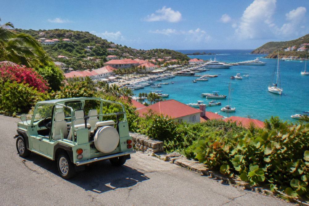 An open-top Mini Moke parked on a narrow road between green hills above the sea