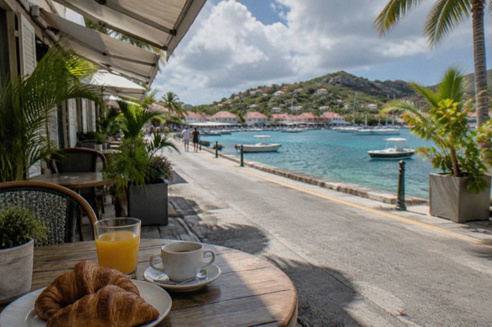 A café table with croissants and orange juice beside a narrow Caribbean street