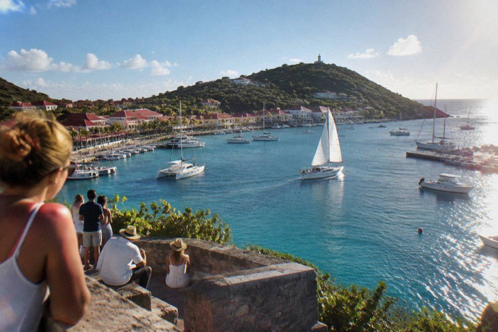 People watching sailboats from a stone rampart above a harbor town