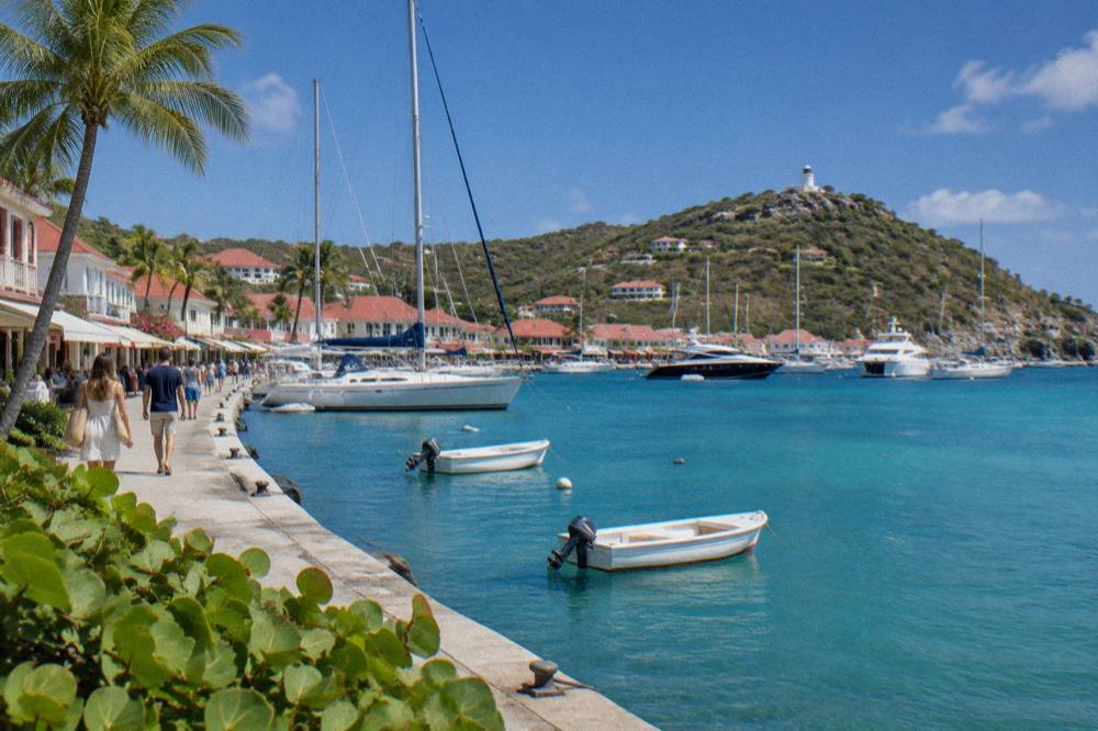 A waterfront street with boutique shopfronts and moored sailboats along a quay