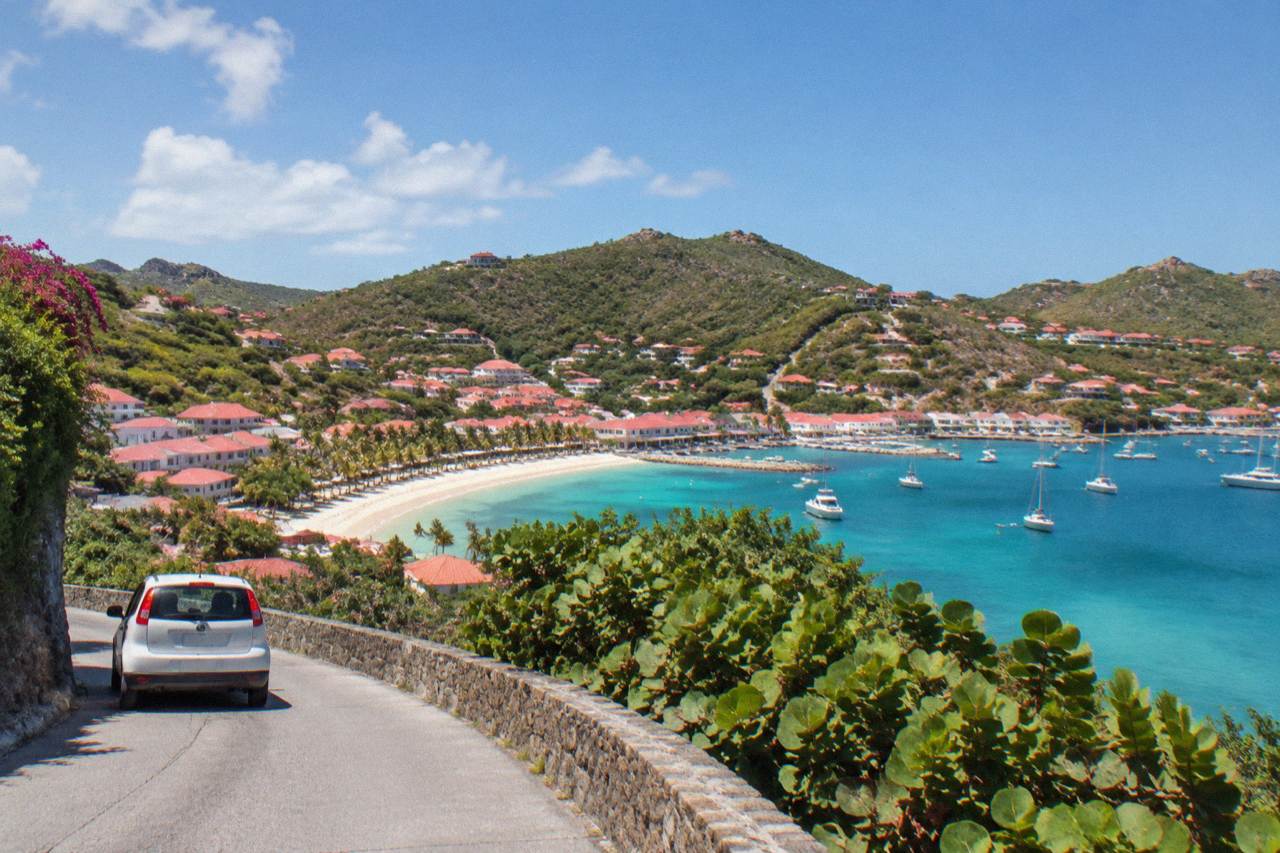 A small rental car on a narrow winding road above a Caribbean bay with red rooftops below