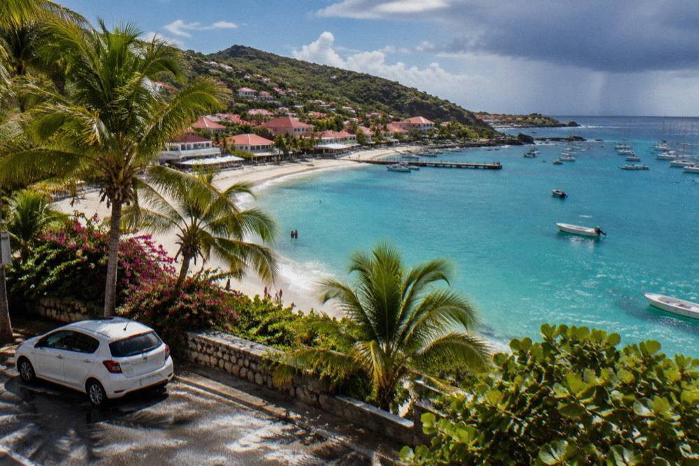 A white compact car parked beside palm trees near a turquoise Caribbean bay