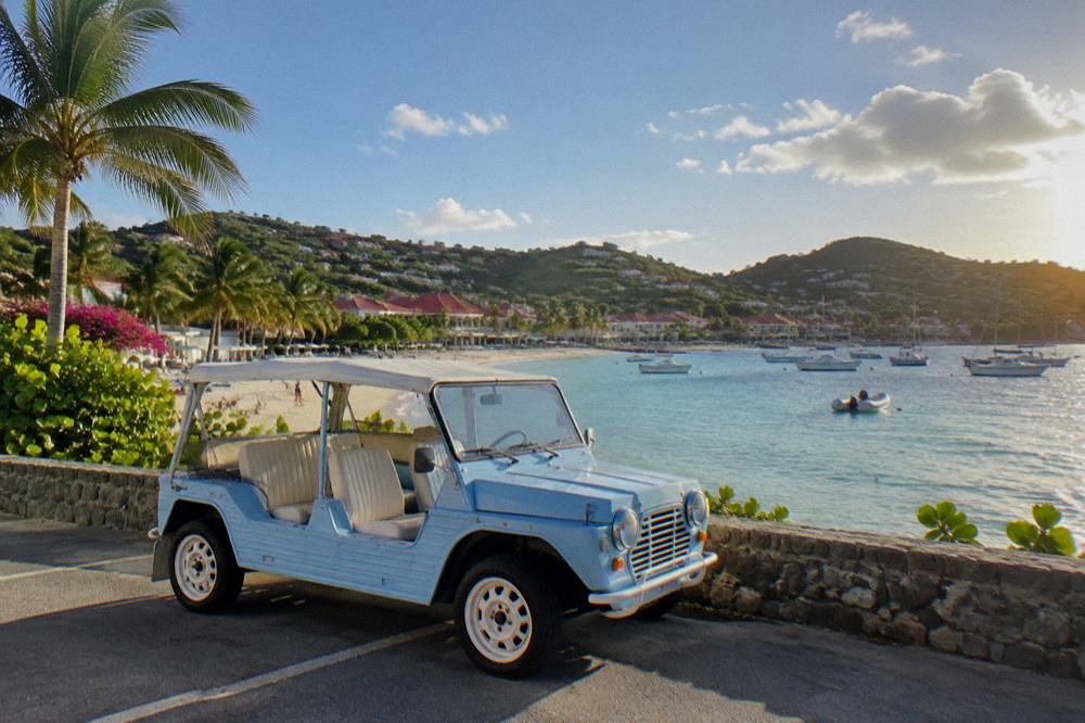 An open-sided Mini Moke parked beside a low stone wall with a beach and palms behind