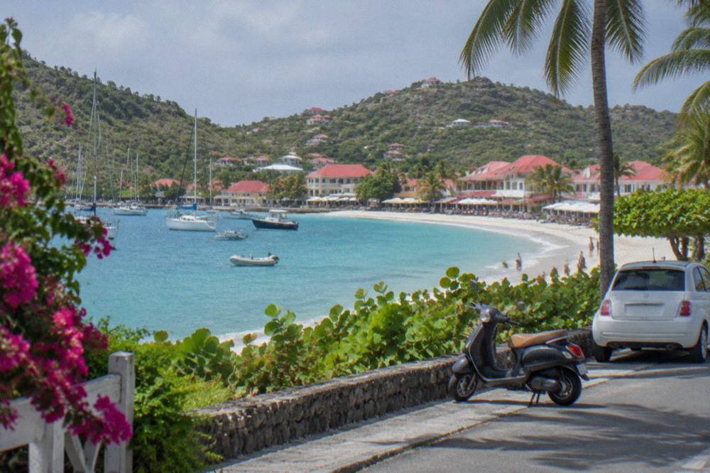 A parked scooter and small car on a waterfront street with yacht masts in a small harbor