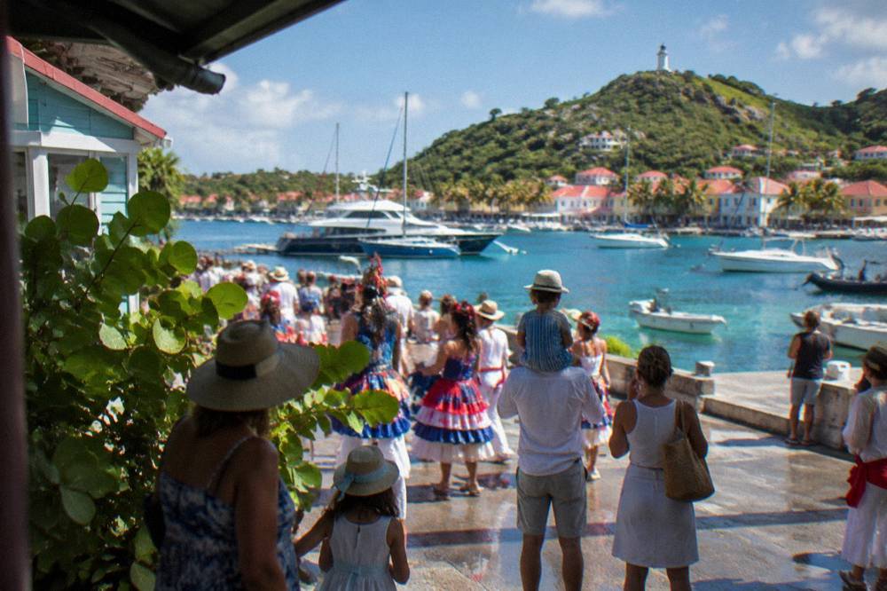 Families in costume watching a parade beside pastel Caribbean shopfronts