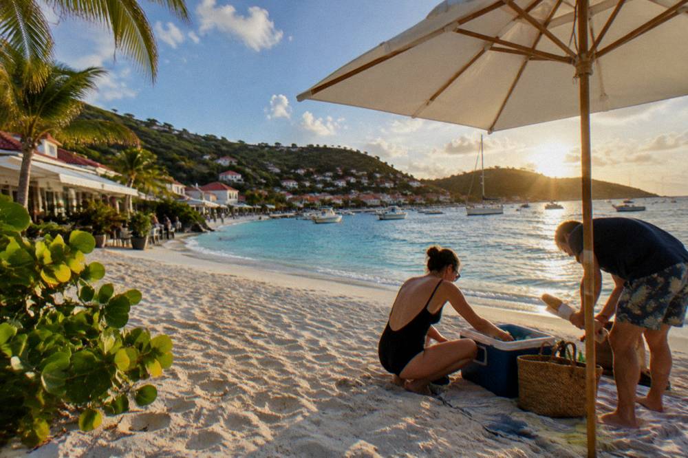 Two people unpacking a cooler and a baguette under a parasol on white sand