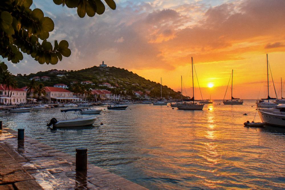 A sunset over Gustavia harbor in St Barts with masts of sailboats silhouetted against an orange sky