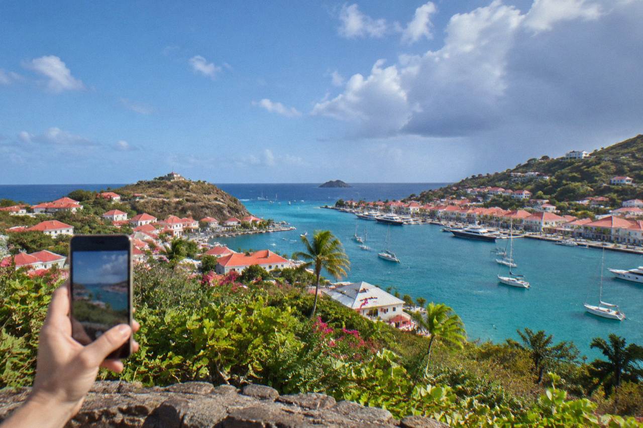 A smartphone held up on a hillside overlooking a Caribbean harbor town and turquoise water