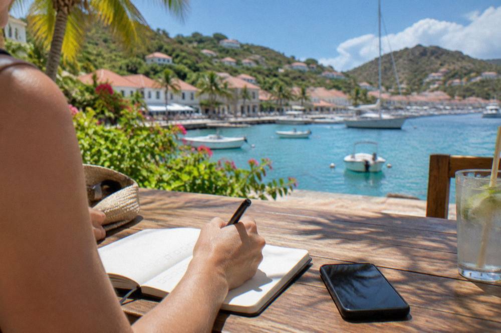 A traveler writing a checklist in a notebook next to a phone on a wooden table