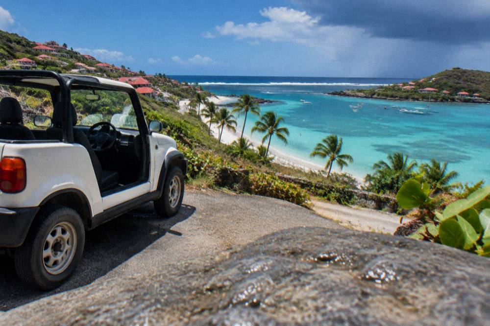 A small open-air rental car parked beside a beach entrance on a hillside road