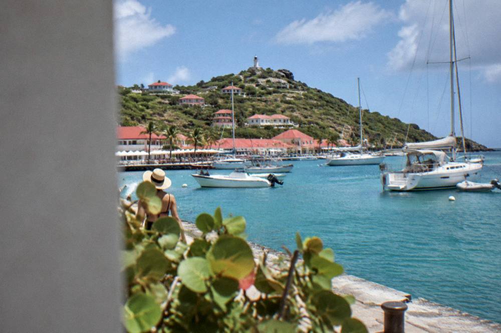 Sailboats anchored in a small harbor beside red-roofed buildings on a green hillside