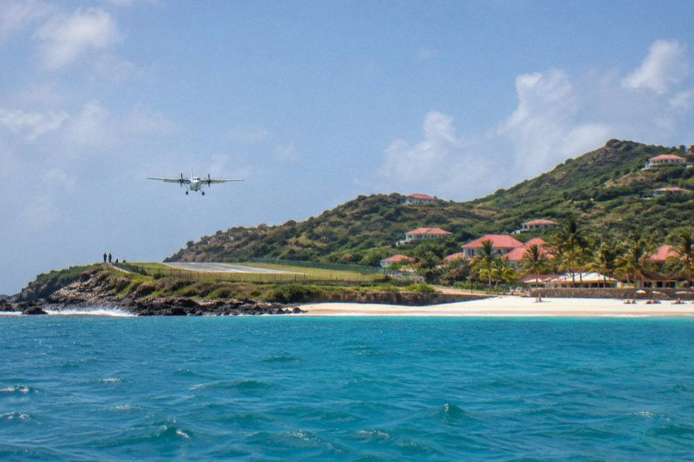 A small turboprop aircraft on a short runway beside a Caribbean beach seen from the water