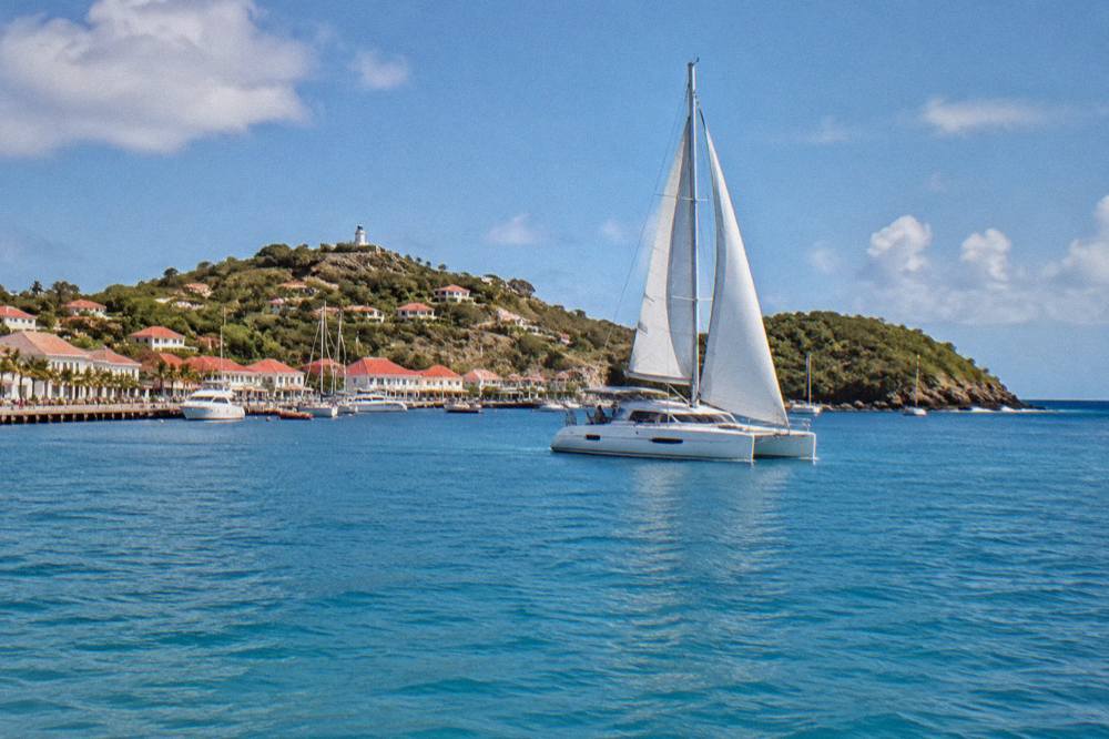 A catamaran under sail on calm blue water near a green island headland