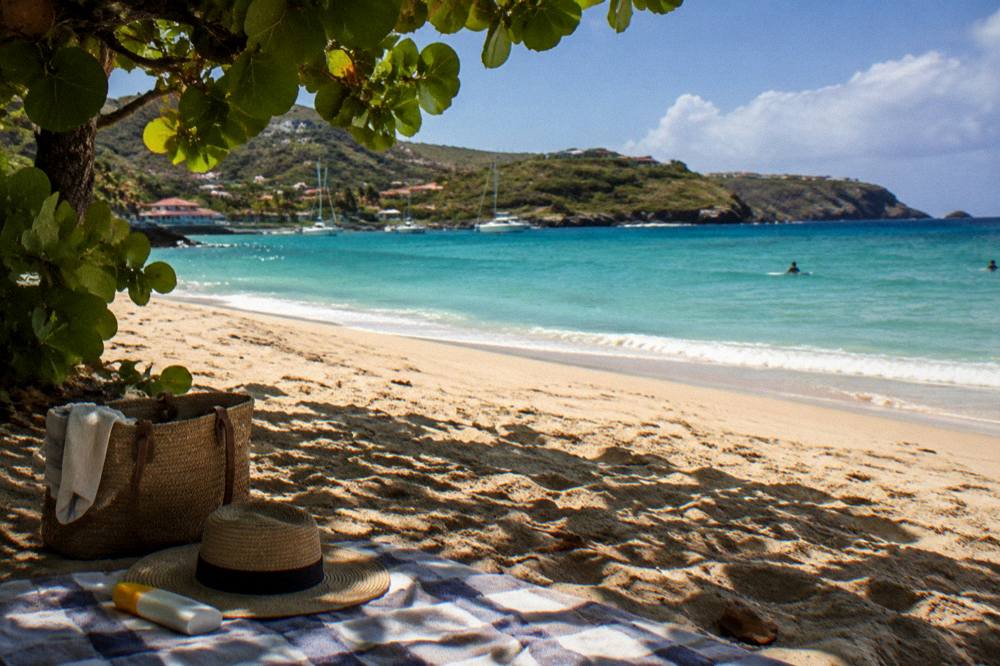 A beach bag, hat and sunscreen on a towel beside turquoise water